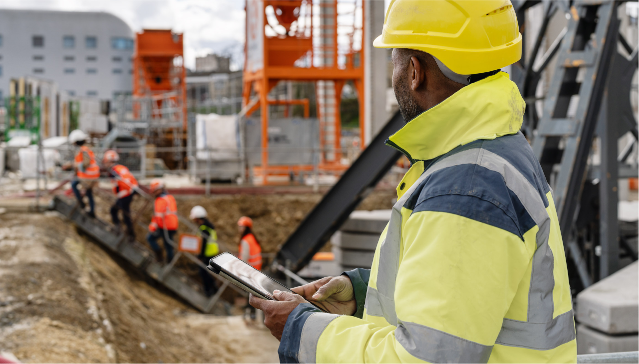 Mann mit Baustellenkleidung und Tablet in der Hand blickt auf Baugrube aus der mehrere Bauarbeiter eine Leiter heraufsteigen.