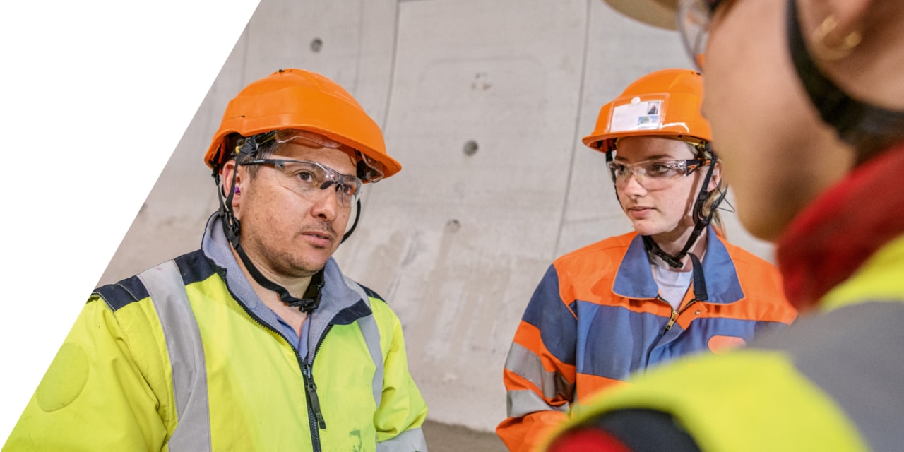 Aufnahme über die Schulter eines Baustellenmitarbeiters. Diese zeigt zwei weitere Personen in Baustellenausrüstung. 