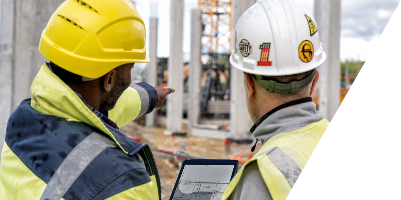 Nahaufnahme zweier Personen in Baustellenbekleidung, deren Blick auf die Baustelle vor ihnen gerichtet ist, während einer von ihnen ein Tablet in der Hand hält. 