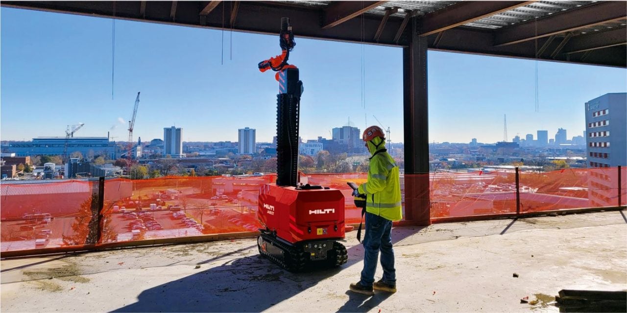 Eine Person in Sicherheitskleidung lenkt einen Baustellenroboter auf einer Baustelle, im Hintergrund die Skyline einer Stadt. 