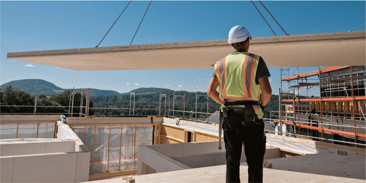 Rückansicht eines Mannes in Sicherheitskleidung, der zusieht, wie ein Haus in Fertigbauweise zusammengesetzt wird. 