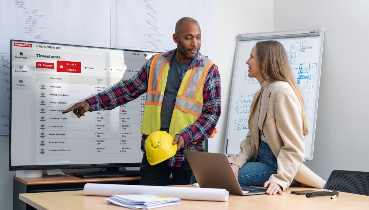 Ein Mann in Arbeitskleidung und eine Frau in Bürokleidung unterhalten sich im Büro vor einem Screen über die Arbeitsverteilung auf der Baustelle.