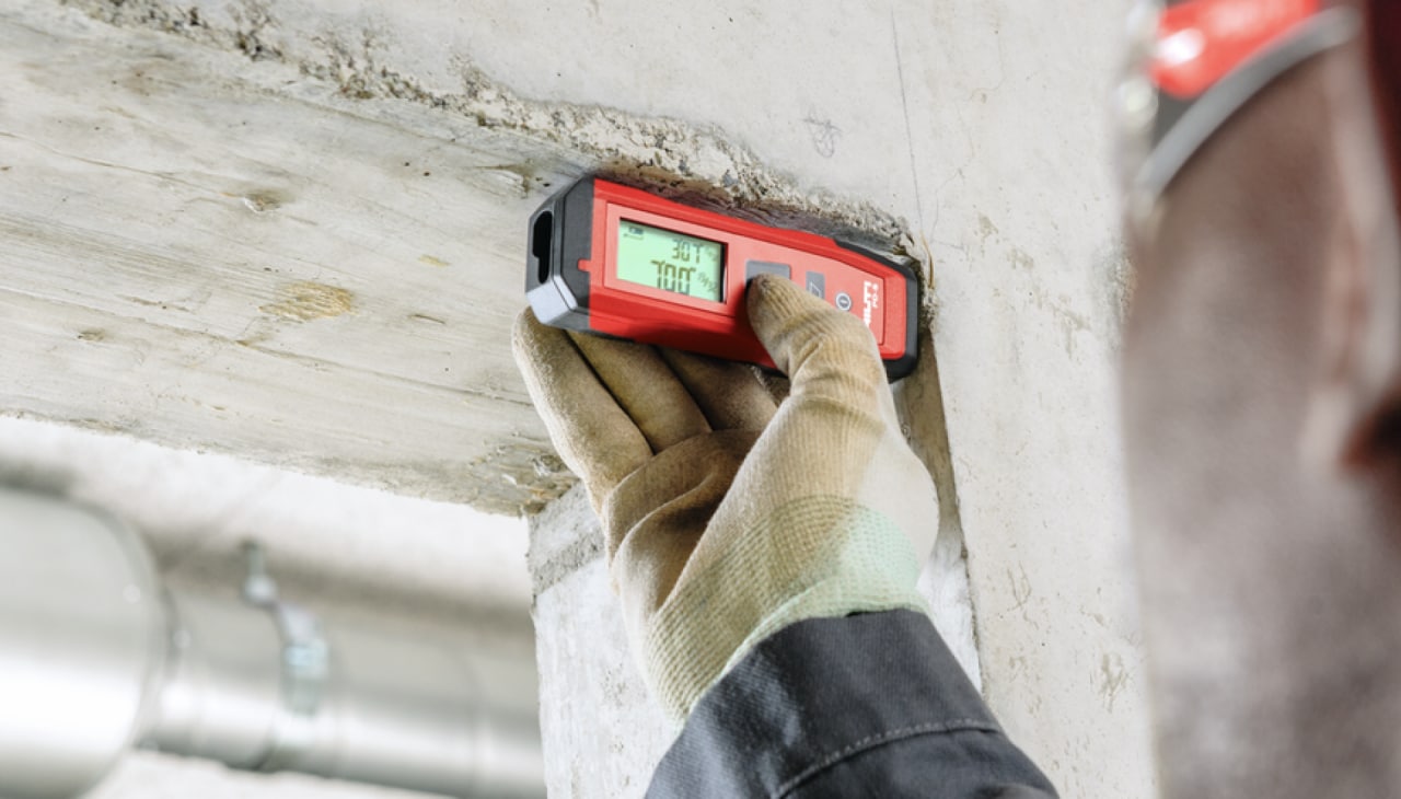 Eine Hand in Arbeitshandschuhen, die mit einem Lasermessgerät Abstände in einem Rohbau prüft.