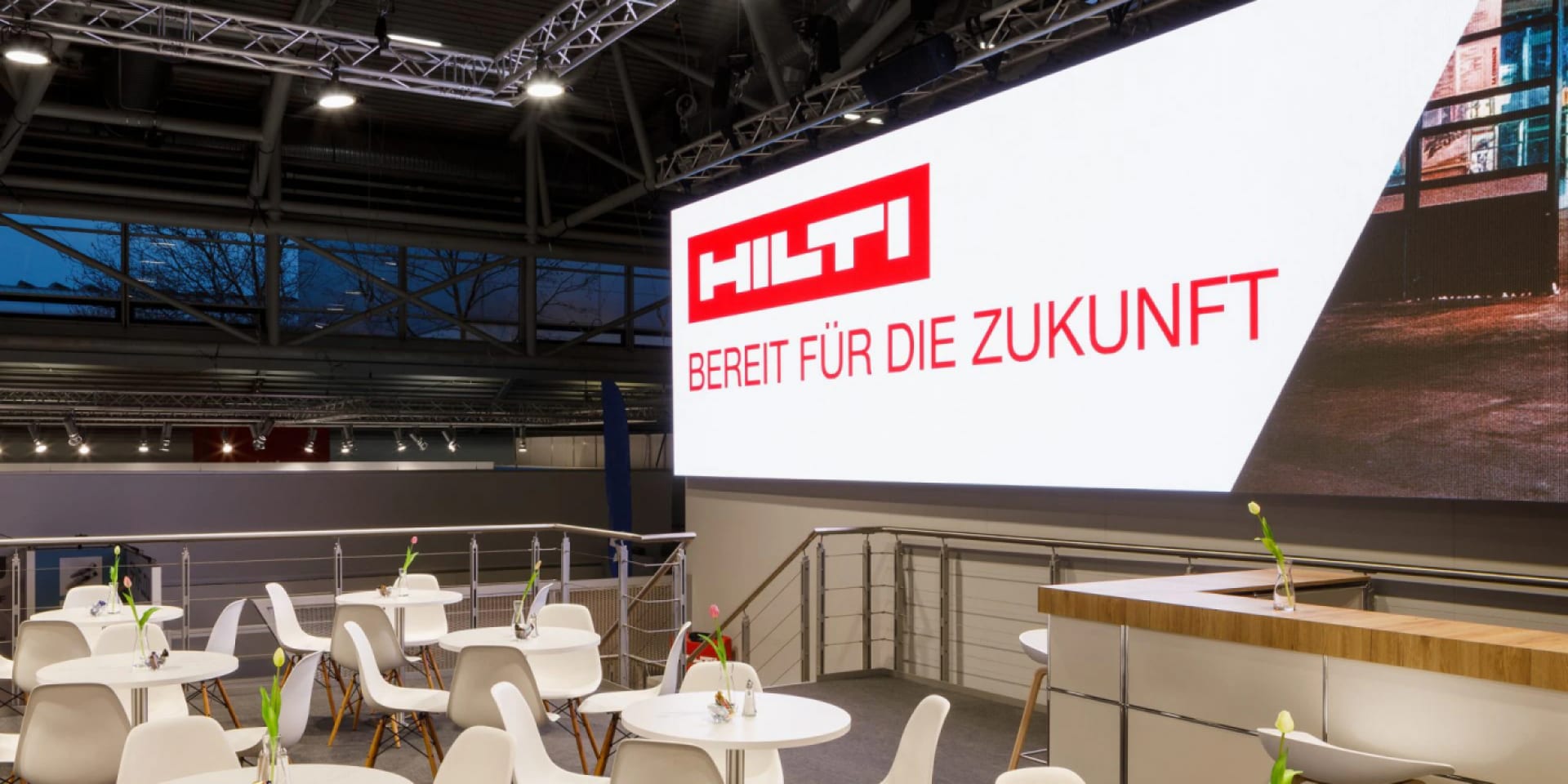 Zu sehen ist eine Besprechungsfläche mit weißen Stühlen und Tischen auf einer Messe. Auf einem Vodeoscreen dahinter sind das Hilti Logo und der Text „Bereit für die Zukunft“ zu sehen.